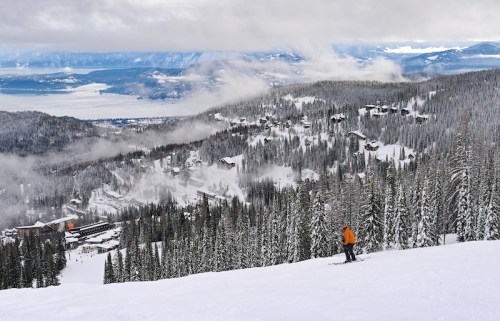 A skier ascends a snowy slope, surrounded by tall pine trees and a scenic view of mountains and a lake in the distance.