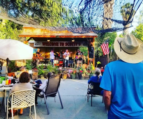 A lively outdoor music performance at a venue, with an audience enjoying the show under a shaded area.