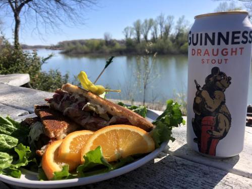 A plate with a sandwich and orange slices, alongside a can of Guinness, with a scenic river view in the background.