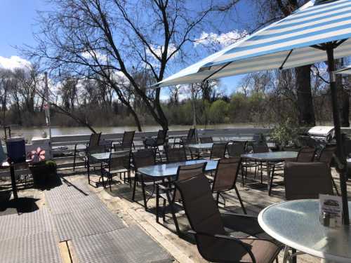 Outdoor seating area by a river, featuring tables and chairs under a striped umbrella, with trees in the background.