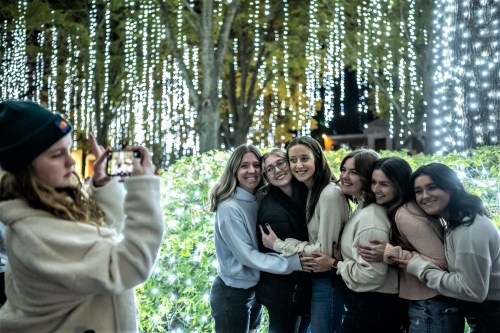 A group of six friends poses together, smiling, in front of a backdrop of twinkling lights while one takes a photo.