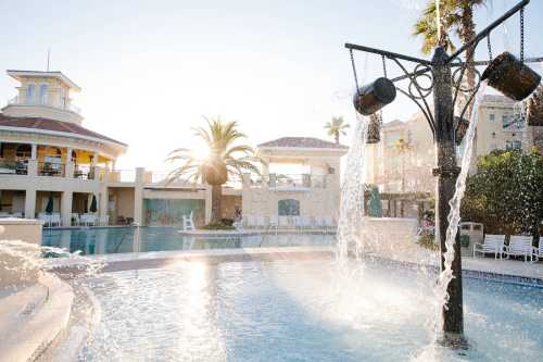 A sunny pool area with a fountain, palm trees, and a resort building in the background.