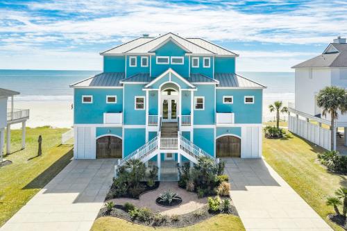 A large blue beach house with a spacious front yard, palm trees, and ocean views under a clear blue sky.