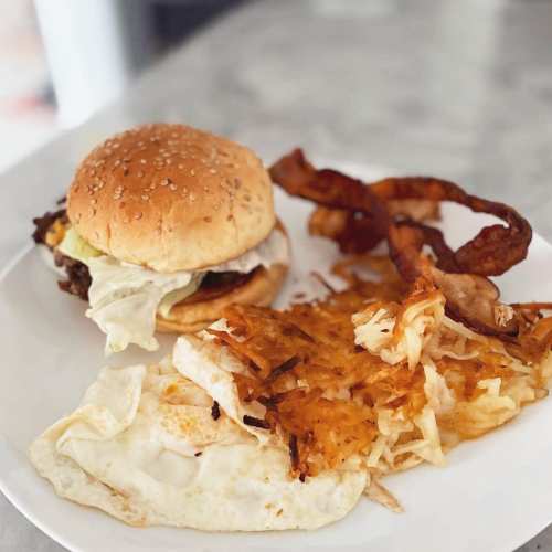 A plate with a sesame seed burger, lettuce, fried eggs, hash browns, and crispy onion rings.