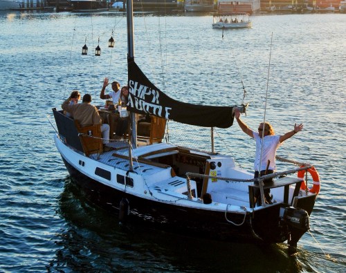 A sailboat on water with people waving, featuring a black sail that reads "SHRIMP ASSULT."
