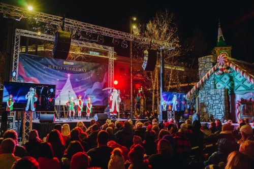 A festive stage with performers in holiday attire, surrounded by an audience and decorated trees at night.