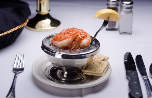A bowl of shrimp topped with sauce and a lemon wedge, served with crackers on a plate, set on a dining table.