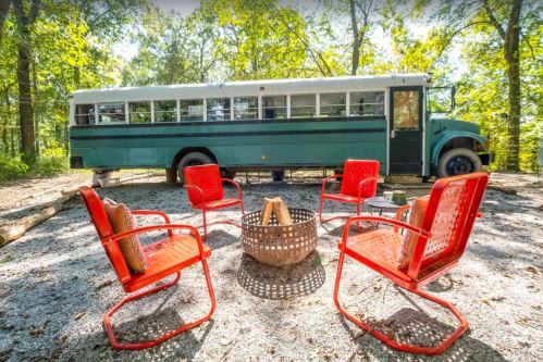 A green converted bus parked in a wooded area, surrounded by red chairs and a fire pit on gravel.