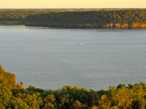 A serene lake scene with a small boat gliding across the water, surrounded by lush greenery and distant hills.