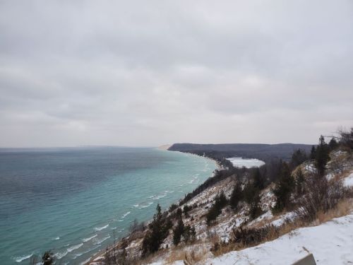 A scenic view of a snowy coastline with turquoise waters and a cloudy sky, featuring rolling hills in the background.