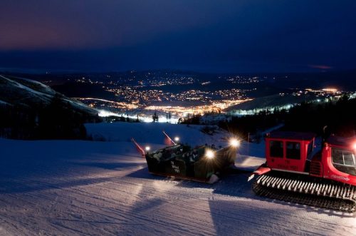 A snowcat vehicle parked on a snowy slope at night, overlooking a lit-up town in the valley below.