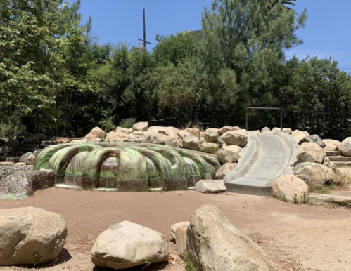 A green, textured play structure surrounded by rocks, with a concrete slide on the right, set in a sunny outdoor area.