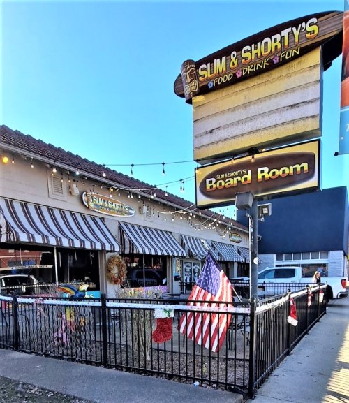 Exterior of Slim & Shorty's, a restaurant with festive decorations and an American flag, featuring a sign for the Board Room.