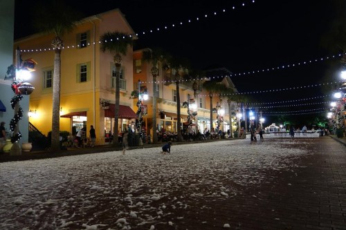 A festive street at night, decorated with lights and palm trees, with a child playing in white snow-like material on the ground.
