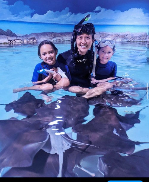 A woman and two girls in wetsuits interact with stingrays in a shallow pool.