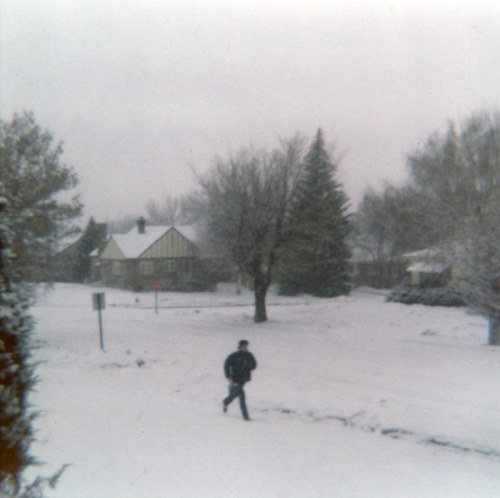 A person walks through a snowy landscape, with trees and a house in the background under a gray sky.