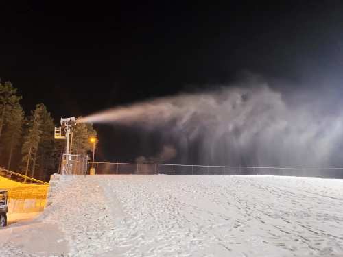 Snowmaking machine spraying artificial snow on a snowy slope at night, with trees in the background.