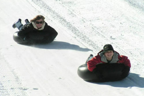 Two people sliding down a snowy hill on black inner tubes, enjoying a winter day.