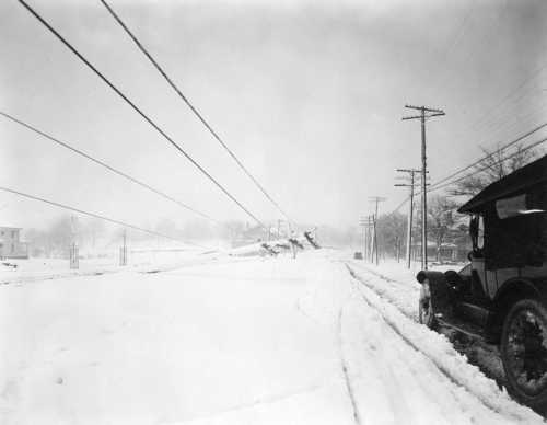 A snowy landscape with a vintage car parked on a road, power lines in the background, and a foggy atmosphere.