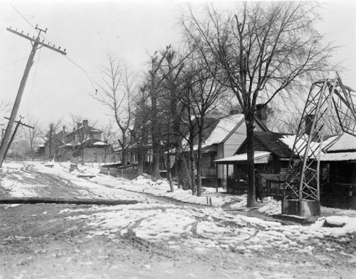 Black and white photo of a snowy street lined with houses and a utility pole, with bare trees and muddy ground.
