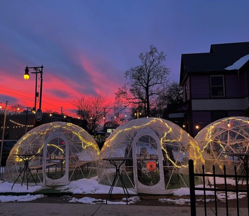 Three illuminated igloos on a snowy ground, set against a vibrant sunset sky with pink and purple hues.