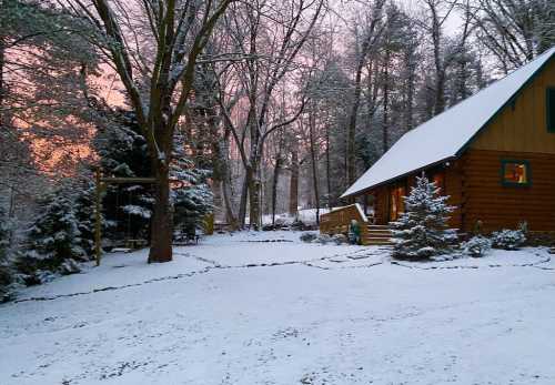 A cozy log cabin surrounded by snow-covered trees at sunset, with a serene winter landscape.