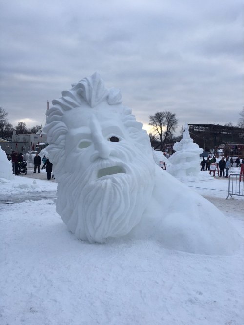 A large snow sculpture of a bearded face, partially buried in snow, with people and trees in the background.