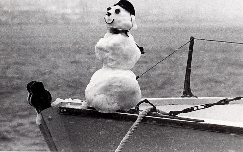 A snowman wearing a hat and scarf sits on the bow of a boat, with a snowy landscape in the background.