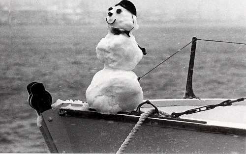 A snowman wearing a hat and scarf sits on the bow of a boat, with a snowy landscape in the background.