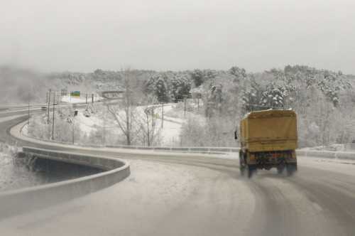 A truck drives on a snowy, winding road surrounded by trees and a winter landscape.