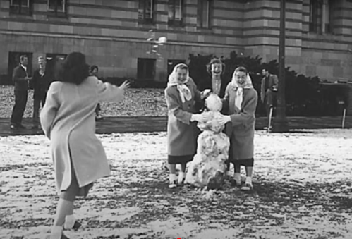 Two girls in winter coats build a snowman while another girl throws a snowball in a snowy park setting.