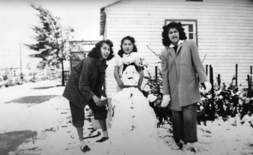 Three women pose with a snowman in a snowy landscape, with a house and trees in the background.