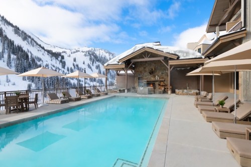 A serene outdoor pool surrounded by snow-covered mountains and lounge chairs, with umbrellas for shade.