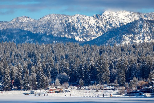 Snow-covered mountains rise above a forest of evergreen trees, with a serene lake and small cabins in the foreground.