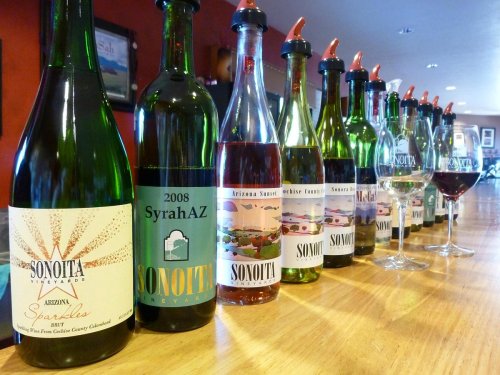 A row of wine bottles from Sonoita Vineyards, with glasses and red wine on a wooden table in a warm setting.
