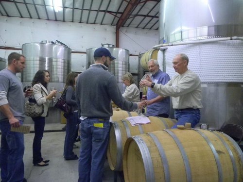 A group of people tasting wine in a winery, interacting with staff near large barrels and stainless steel tanks.
