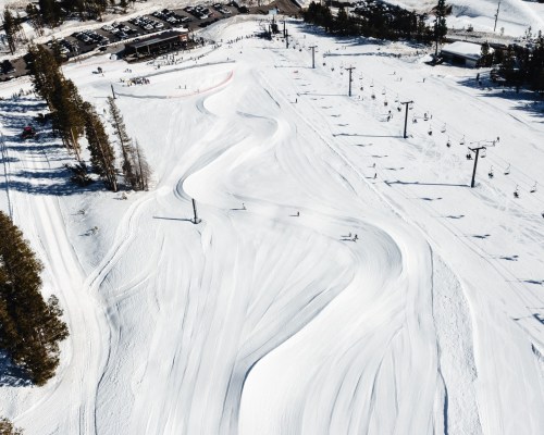 Aerial view of a snowy ski slope with winding trails, ski lifts, and a parking area in the background.
