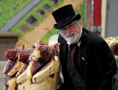 A man in a top hat and formal attire smiles beside wooden bear carvings at an event.