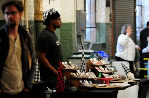 A vendor stands at a market booth displaying handmade items, while a customer browses nearby.