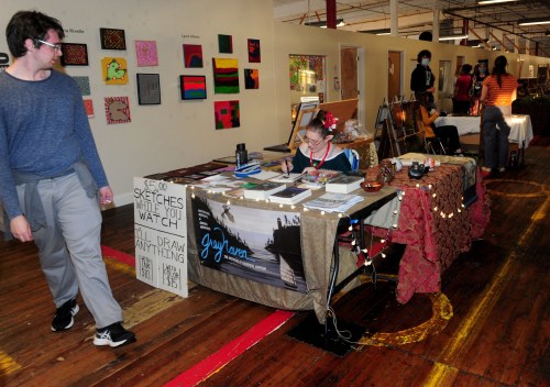 A person walks past an art booth with sketches and colorful artwork displayed on the walls in a spacious venue.