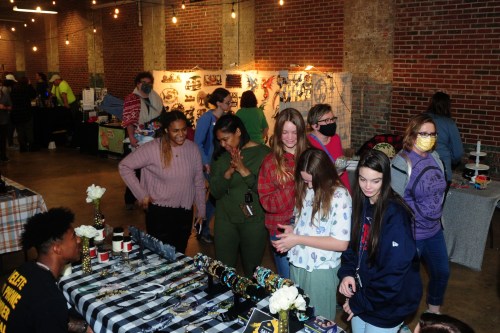 A group of people browsing a market with handmade goods, surrounded by colorful displays and festive lighting.