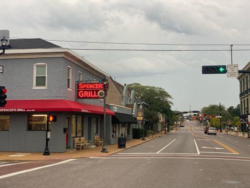 A street view featuring Spencer's Grill with a neon sign, empty sidewalks, and traffic lights under a cloudy sky.