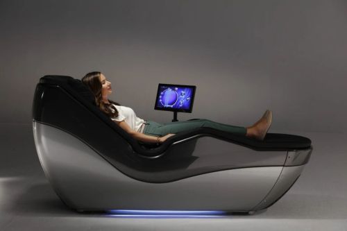 A woman relaxes in a modern massage chair with a screen displaying a graphic, set against a gray background.