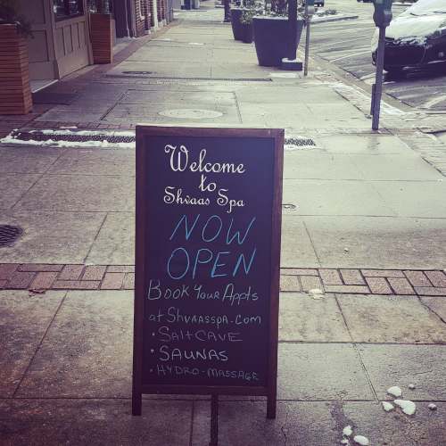 A sidewalk sign for Shivasa Spa, announcing it's now open and offering services like salt cave and sauna.