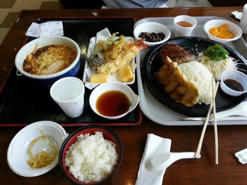 A tray with a variety of Asian dishes, including noodles, tempura, rice, and dumplings, alongside small bowls of sauces.