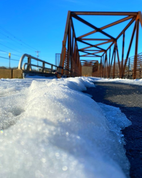A snowy path leads to a metal bridge under a clear blue sky.