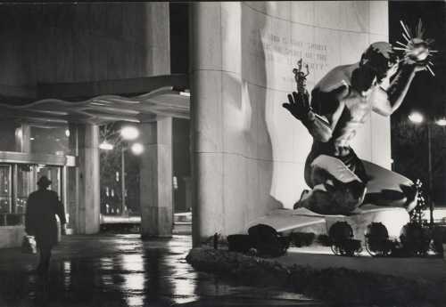 A man walks past a large statue of a kneeling figure holding a torch, illuminated at night near a building entrance.