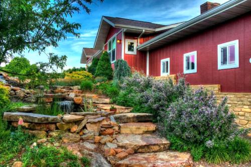 A red house with large windows, surrounded by lush greenery and a stone pathway leading to a small pond.