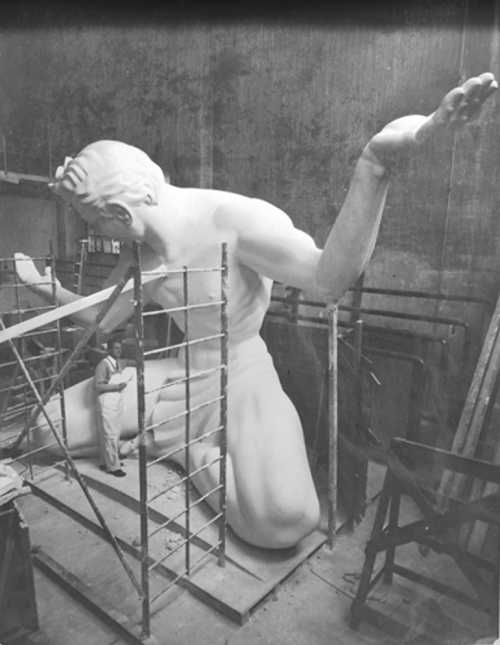 A large statue of a kneeling figure is being worked on in a workshop, with a person standing nearby on scaffolding.