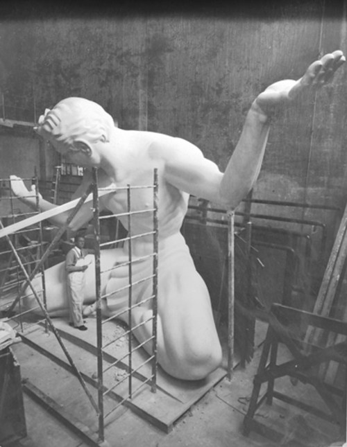 A large statue of a kneeling figure is being worked on in a workshop, with a person standing nearby on scaffolding.